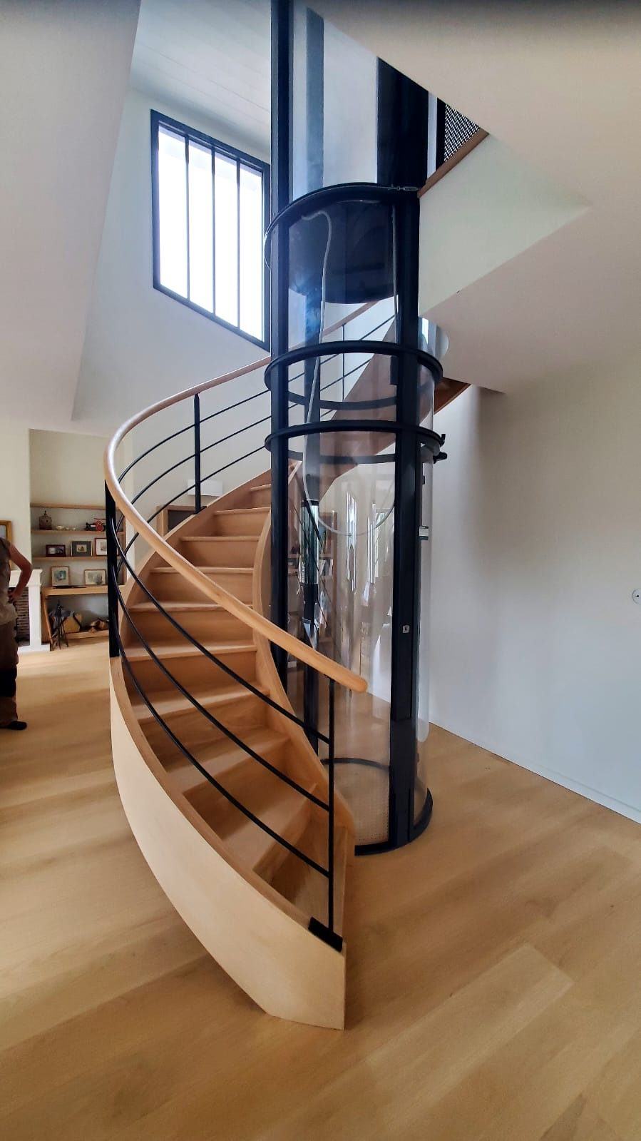 Escalier cintré en bois autour d’une gaine d’ascenseur vitrée